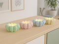 Four pastel-colored pumpkin-shaped candles on a wooden surface with a blurred indoor background.