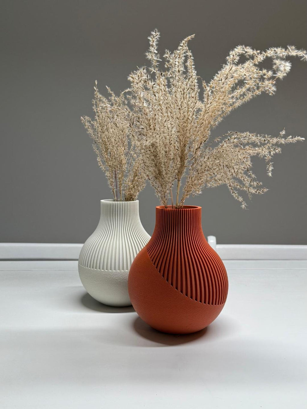Two ceramic vases with textured designs, one white and one terracotta, containing dried plants on a neutral background.